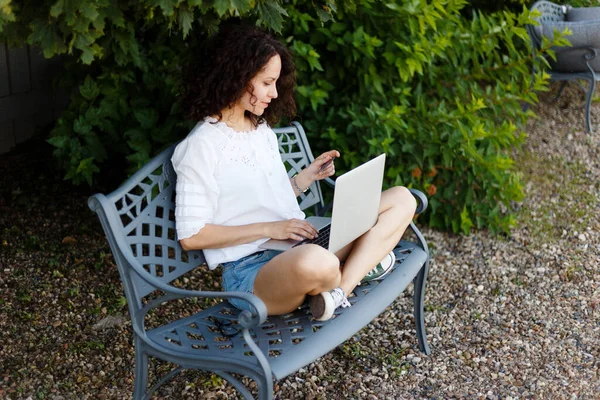 一位年轻女子双腿交叉坐在公园的长椅上 她用笔记本电脑和信用卡在网上购物 — 图库照片