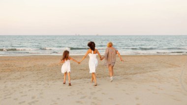 İki yetişkinin ve gün batımında kumsalda koşan küçük tatlı bir kızın arka görüntüsü. Yatay görünüm.