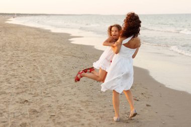 Yatay görüş. Anne, kızını sahilde kucaklıyor. Tatilde ve seyahatte mutlu bir aile. Anne sahilde kızını kucaklıyor ve oynuyor..