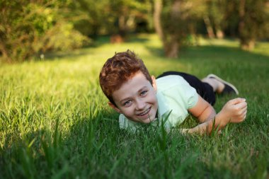 Gülümseyen genç bir çocuk yeşil çimlerde yuvarlanıyor, parkın tadını çıkarıyor, güneşli bir yaz gününde kameraya bakıyor..