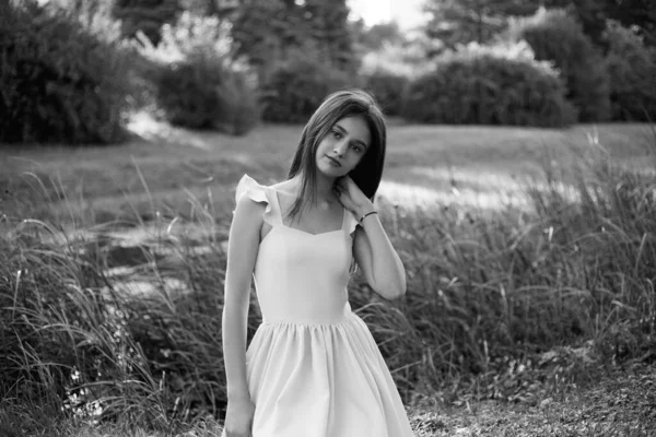 Frontal portrait of a beautiful young girl dressed in dress seated on grass, touching her neck, looking at camera. — Stock Photo, Image