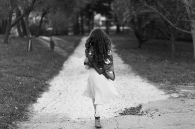 Siyah-beyaz fotoğraf. Ormandaki köy yolunda yürüyen, elbiseli bir kızın arka görüntüsü..