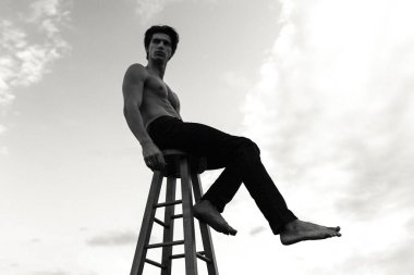 Yatay görüş. Dışarıda poz veren yakışıklı, üstsüz, kaslı genç adam. Fotoğraf beyaz ve siyah. Koltukta oturan erkek model, gökyüzü arka planında.