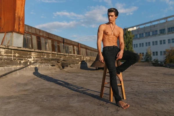 Handsome sexy and fit sportive young man with naked torso, dressed in black jeans, posing on old abandoned factory. Horizontal view.