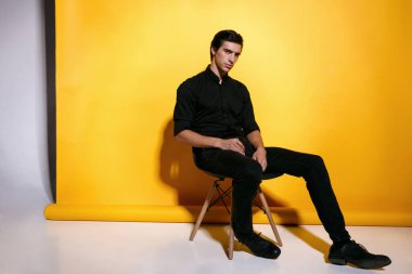 Sarı arka planda oturan, siyah elbiseli, kendine güvenen yakışıklı bir gencin portresi. Yatay görünüm.