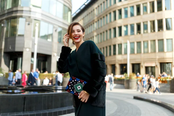 Close up portrait of a smiling woman walking down the street. Fashionable girl wearing dark green fur coat and clutch.