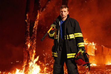Hüzünlü teçhizatlı itfaiyeci, kaskı elinde tutuyor, bu da alevler içindeki bir ormanın arka planında duruyor. Yatay görünüm.