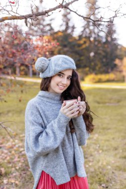 Gülümseyen güzel esmer bir kızın ön portresi şık, yumuşak giysiler içinde, sonbahar parkında sonbahar yeşillikleri altında bir bardak tutuyor. Golden Autumn Park arka planı. Dikey görünüm.