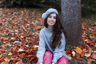 Gri bereli ve sonbahar elbiseli genç kız sonbahar mevsiminde bir ağacın yanındaki yaprakların üzerinde oturuyor..