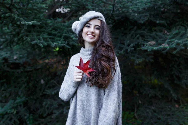Gri elbiseli gülümseyen kız elinde kırmızı bir yaprak, köknar ağacının üzerinde, sonbahar arka planında, dışarıda çekilmiş. Yatay görünüm.