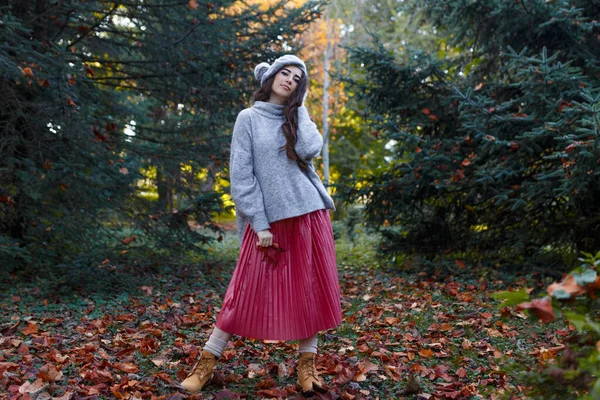 Gri elbiseli, kırmızı etekli neşeli kız elinde kırmızı bir yaprak, köknar ağacının üstünde, sonbahar arka planında, dışarıda çekilmiş..