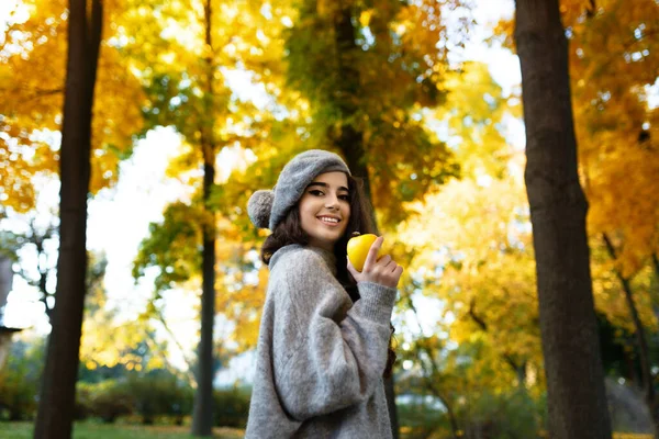 Gri bereli ve kazaklı güzel bir kız, güzel bir makyajı var, elinde sarı bir elmayla, güneşli bir günde. Yatay görünüm.