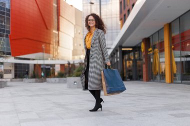 Paltolu, şık giyinmiş, alışveriş çantalı, binaların arkasında oturan neşeli, kıvırcık bir genç kadın resmi..