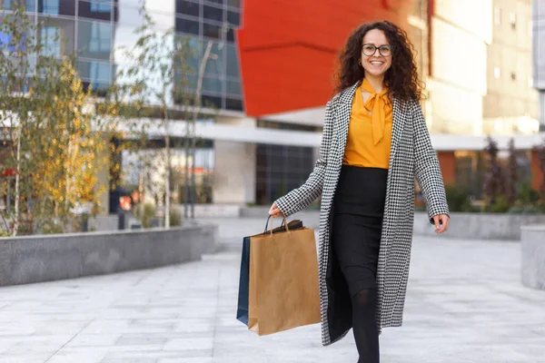 Outdoors portrait of a beautiful curly young woman with many shopping bags, over buildings background. Horizontal view. Copy space.
