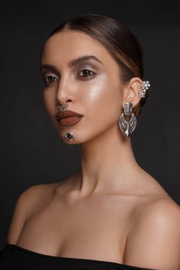 Alçak kesim siyah elbiseli, makyajlı, gümüş bijouterie 'li, gri arka planda kameraya bakan şehvetli kadın..