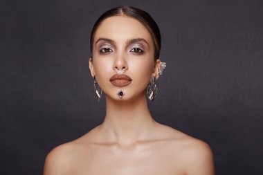 Göz kamaştırıcı bir kadın model, makyaj mücevherleri, kameraya ön tarafı bakıyor, stüdyoda poz veriyor, koyu gri arka planı var..