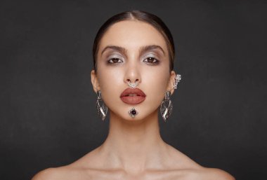 Yatay görüş. Göz kamaştırıcı bir kadın model, makyaj mücevherleri, kameraya ön tarafı bakıyor, stüdyoda poz veriyor, koyu gri arka planı var..