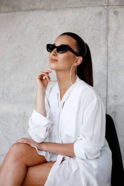 Yuvarlak küpeli, at kuyruklu, siyah gözlüklü, beyaz gömlekli, beton arka planda..