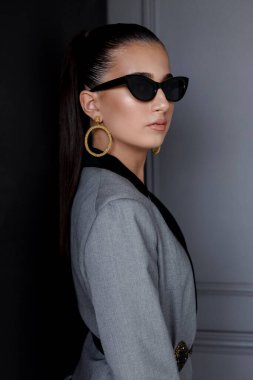 Dikey görünüm. Şık takım elbiseli bir kızın portresi ve siyah güneş gözlüklü, saçları gri duvar arkasında toplanmış..