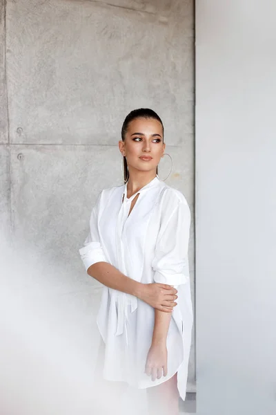 Carina ragazza bruna in elegante camicia lunga bianca, con i capelli raccolti in una coda di cavallo, isolata su un muro di cemento. — Foto Stock