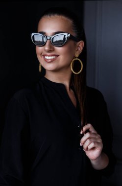 Zarif siyah gömlek ve güneş gözlüklü, makyajlı, koyu arkaplan üzerine gülümseyen güzel bir kızın portresi..