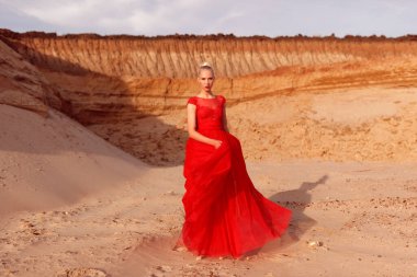 Kumların arka planında uçan kumaşlı kırmızı elbiseli şehvetli genç bir kadının tam boy görüntüsü..