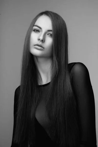 Frontal portrait of a attractive young woman with long hair and make-up, wear in black bodysuit, looking at camera, over grey background. Black and white photo.