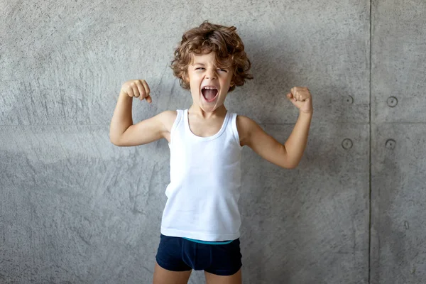 Kıvırcık saçlı, küçük cesur bir çocuğun ön portresi. Yumruklarını havada tutuyor ve arka planda beton gri bisepsleri gösteriyor..