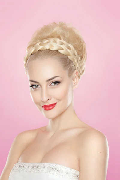 Frontal portrait of a cute blonde young woman with bright make-up and hairstyle, with bare schoulders, over light pink background.