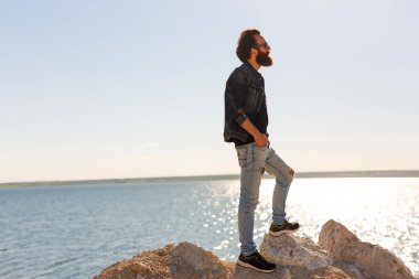 Güneş gözlüklü şık sakallı hippi bir çocuğun dünya turu boyunca sakin bir denizin yanında poz verişi. Yandan ateş et..