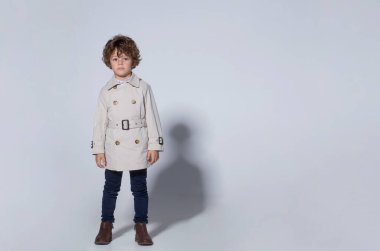 Şık kıvırcık bir çocuk, bej bir siperde giyinmiş, stüdyoda poz veriyor, beyaz arka planda kameraya bakıyor. Boşluğu kopyala.