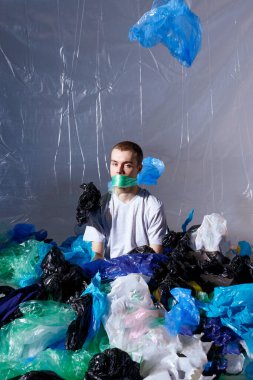 Plastik poşetlerle çevrili, ağzı plastik bir torbayla bağlanmış oturan bir adam kameraya bakıyordu..