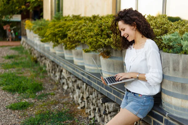 Profilporträtt av leende lockig kvinna i avslappnade kläder som håller och arbetar på sin bärbara dator utanför. Horisontell vy. — Stockfoto