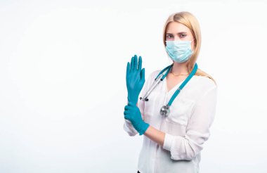 Tıbbi maskeli genç bir kadın koluna koruyucu steril eldiven takıyor. Gri arka planda izole edilmiş. Koronovirüsten korunuyor.