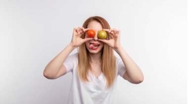 Gri arka planda duran ve gözlerini domatesle kapatan mutlu genç kadın.