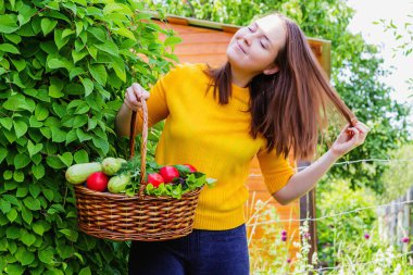 Genç bir esmer kadın, üzerinde domates, bitki ve kabak hasadı bulunan hasır bir sepetle bahçede parlak sarı bir ceketle duruyor.