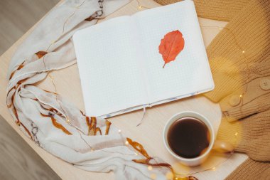 Boş sayfalarda açık günlük bir günlük, bir fincan sıcak kahve, düğmeli bej bir hırka, ahşap bir masanın üzerinde sarmal bir şekilde duran bir çelenk. Kırmızı yapraklar bir kazağın, not defterinin ve masanın üzerinde. Üst görünüm.