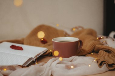 Boş sayfalarda açık günlük bir günlük, bir fincan sıcak kahve, düğmeli bej bir hırka, ahşap bir masanın üzerinde sarmal bir şekilde duran bir çelenk. Kırmızı yapraklar bir kazağın, not defterinin ve masanın üzerinde. Yan görünüm.