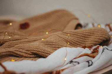 Masanın üzerinde bej renkli yün örgü bir kazak ve açık kahverengi bir pamuk atkı var. Tepede bir çelenk var. yan görünüm