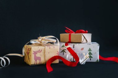Siyah arka planda, ambalaj kağıdına sarılı ve kurdelelerle bağlanmış Noel hediyeleri, kırmızı kurdeleyle bağlanmış küçük bir el işi kutusu, beyaz kurdeleyle bağlanmış büyük bir kutu var.