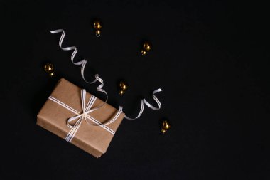 Geri dönüşümlü el sanatları kağıdına sarılı hediye kutusu ve siyah arka planda beyaz ve altın bir kurdele ile bağlanmış, ağaçtaki Altın Noel toplarının yanında. Tatil için sürpriz..