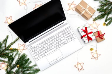 Noel alışverişi. Beyaz arka planda açık bir laptop, kırmızı kurdeleli hediye kutuları, yeşil köknar dalları, yıldız çelenkleri. Kış tatilinde online ticaret.
