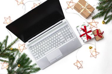 Noel alışverişi. Beyaz arka planda açık bir laptop, kırmızı kurdeleli hediye kutuları, yeşil köknar dalları, yıldız çelenkleri. Kış tatilinde online ticaret.