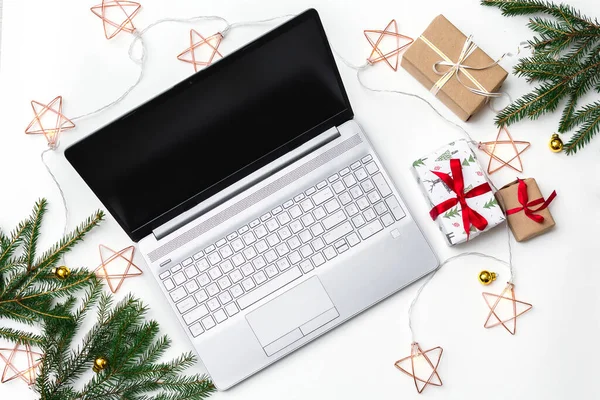 Noel alışverişi. Beyaz arka planda açık bir laptop, kırmızı kurdeleli hediye kutuları, yeşil köknar dalları, yıldız çelenkleri. Kış tatilinde online ticaret.