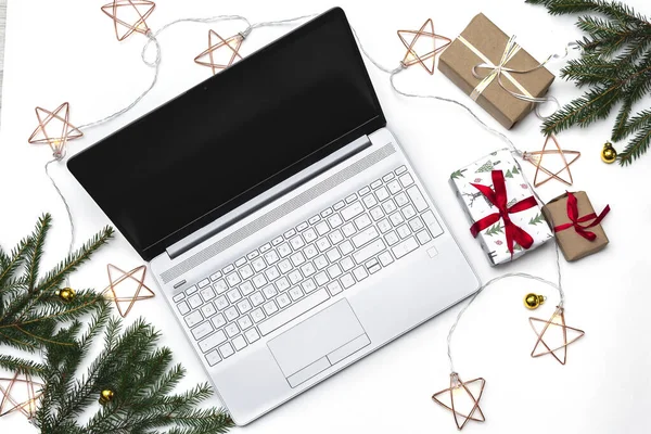 Noel alışverişi. Beyaz arka planda açık bir laptop, kırmızı kurdeleli hediye kutuları, yeşil köknar dalları, yıldız çelenkleri. Kış tatilinde online ticaret.