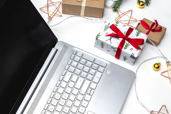 Noel alışverişi. Beyaz arka planda açık bir laptop, kırmızı kurdeleli hediye kutuları, yeşil köknar dalları, yıldız çelenkleri. Kış tatilinde online ticaret.