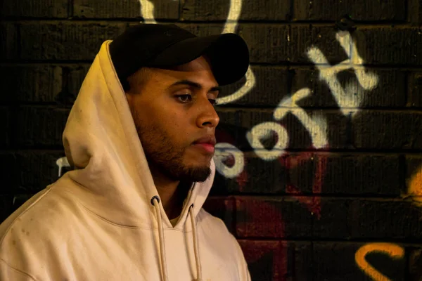 Parlayan sarı ışığın altında poz veren bir adam. Duvarın yanında grafiti sanatıyla dolu bir duvar.