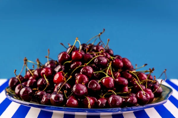 Mavi ve beyaz çizgili bir masada, büyük bir toprak kabında taze, kestane rengi kirazlar..