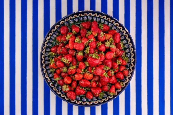 Mavi ve beyaz çizgili arka planda olgun taze çilekleri olan büyük bir toprak kaplama tabağı..