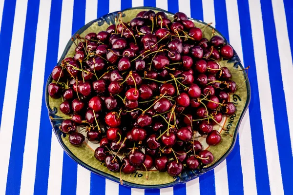 Mavi ve beyaz çizgili arka planda olgun kestane rengi kirazlarla dolu büyük bir toprak kaplama tabağı..